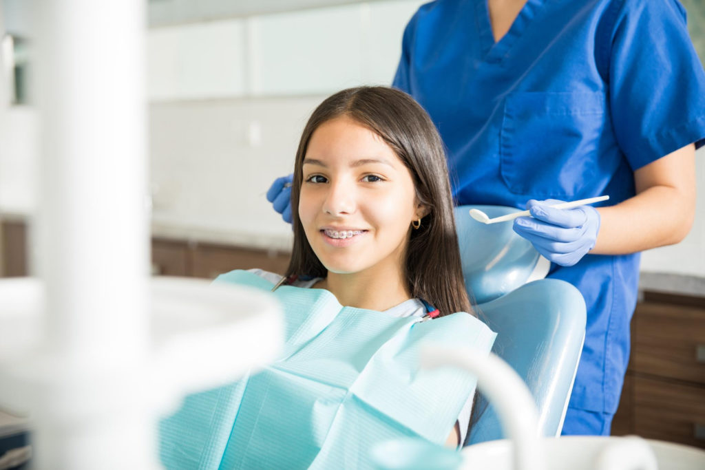 portrait of smiling teenage girl with braces sitting on chair while dentist standing in clinic explaining how do braces change your face shape