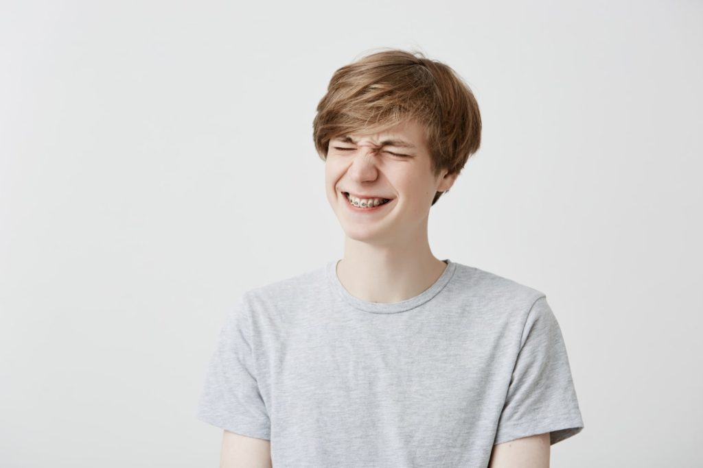 cheerful young man has overjoyed expression clenches teeth with braces in gray t shirt discovering how long do your teeth hurt after getting braces