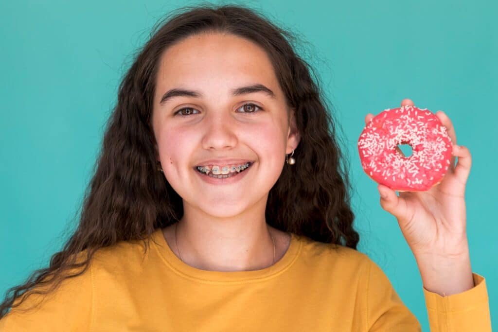 Smiling girl with braces showing a glazed donut and happy to know how long after getting braces can you eat solid food