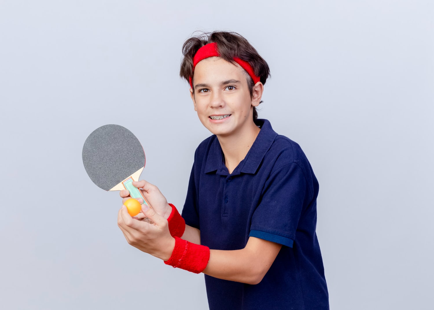 smiling young sporty boy wearing headband wristbands with dental braces holding ping pong racket ball questioning can i play sports with braces
