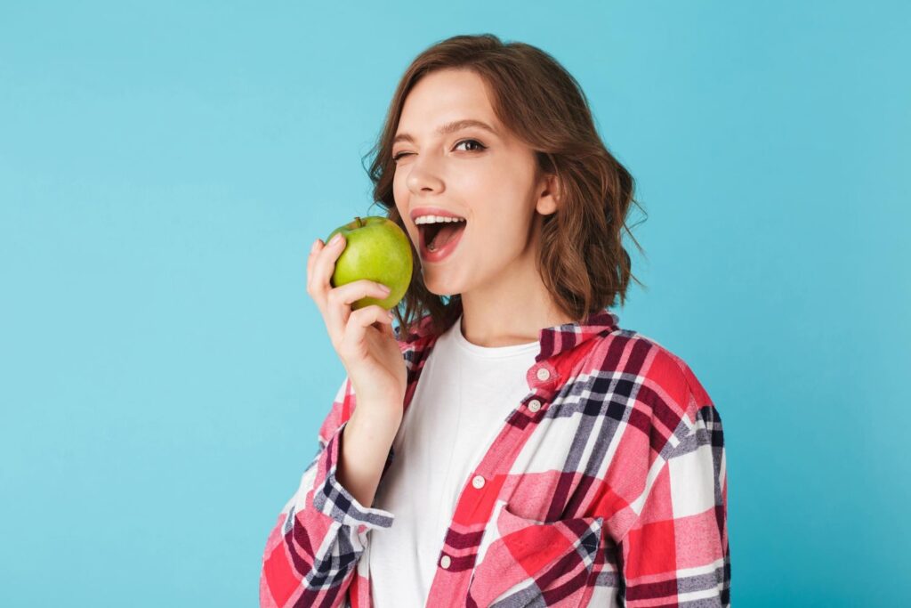 Young woman in a checkered shirt eating a green apple, looking happily at the camera against a blue background, explaining if you can eat after getting a dental crown