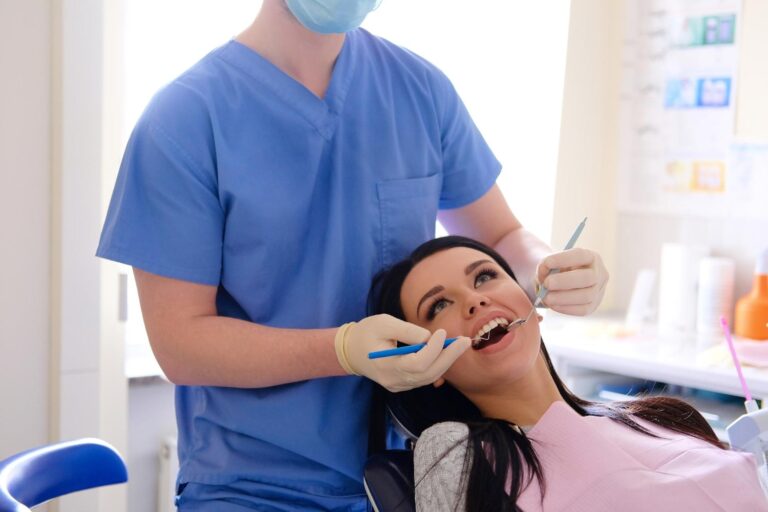 Dentist in blue uniform wearing white gloves attending brunette caucasian woman looking at her mouth with a small mirror